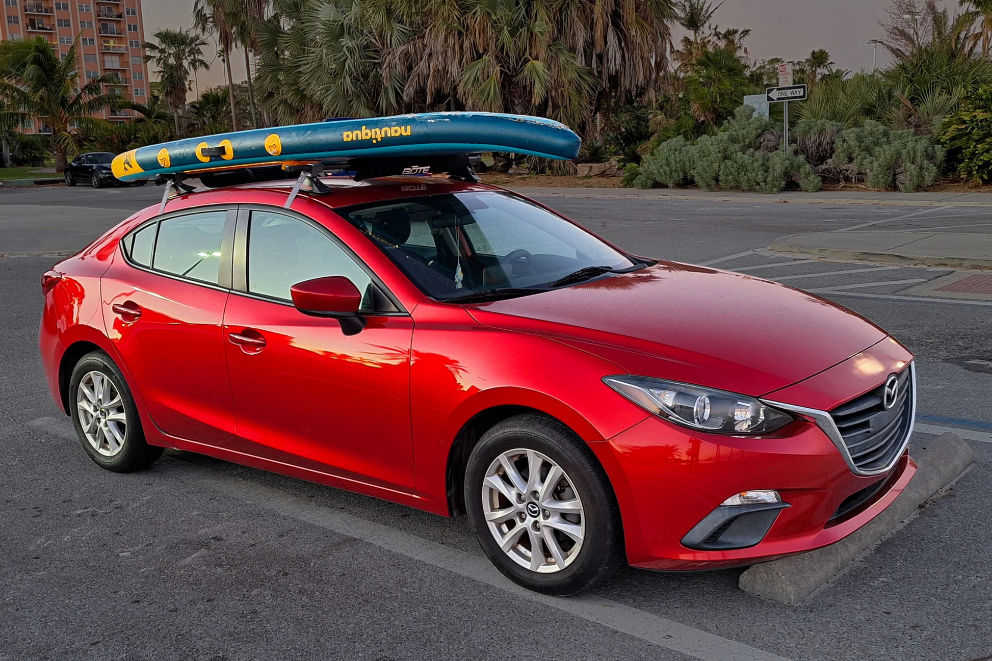 Red car with a surfboard on top in a parking lot with palm trees in the background