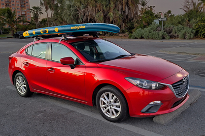 Red car with a surfboard on top in a parking lot with palm trees in the background